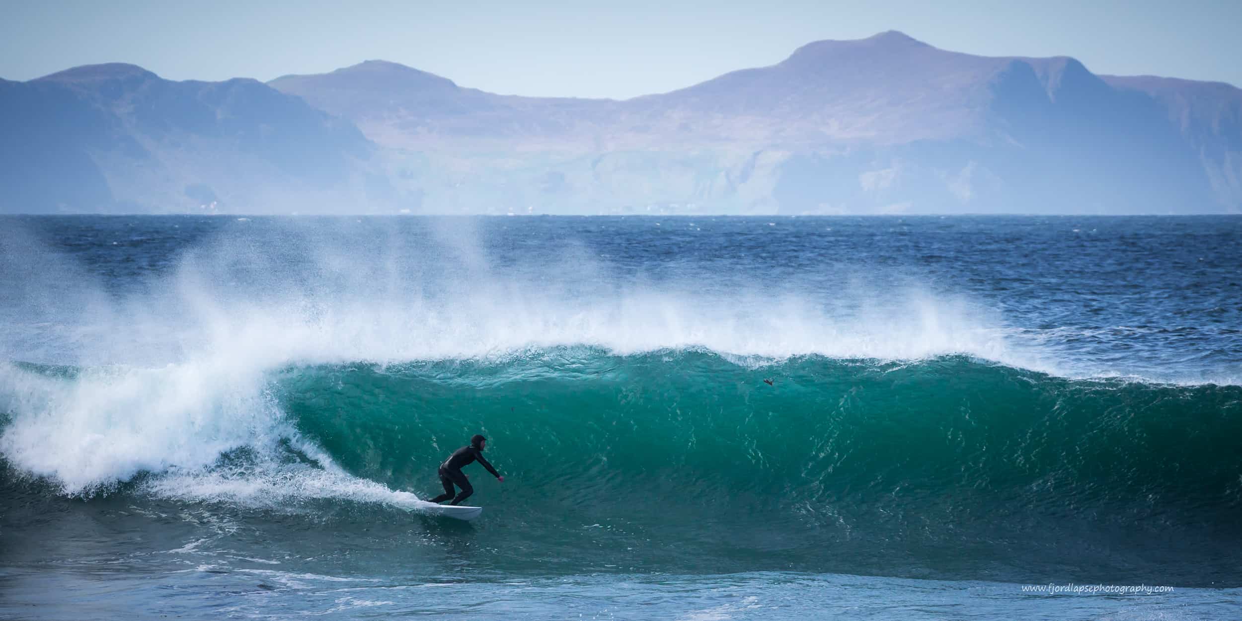 Surfer bølge på Sunnmøre. Fjell i bakgrunnen.