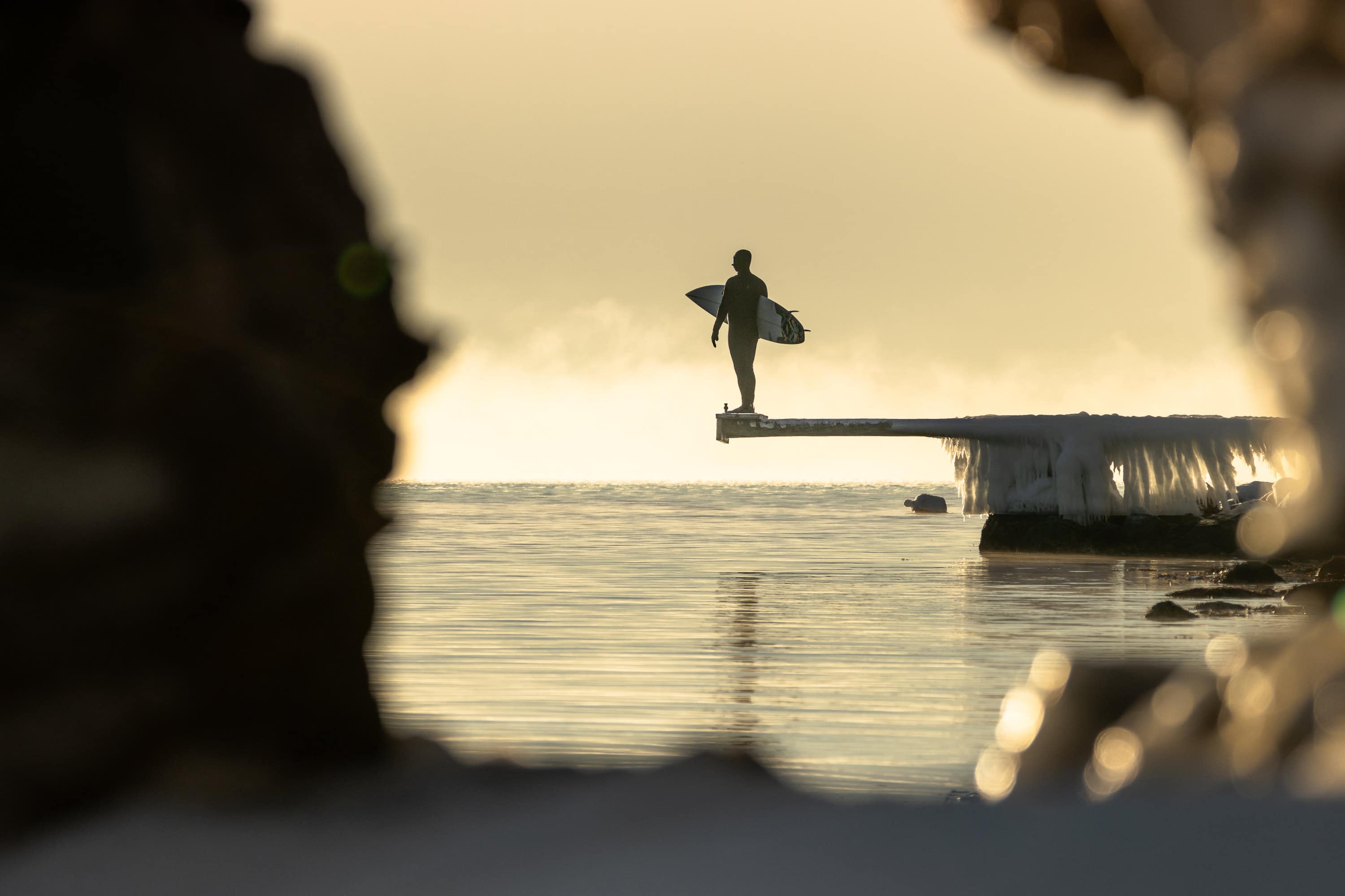 Surfer står på frossen brygge og ser utover havet.